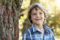 Счастливый стильный маленький мальчик в шортах и клетчатой рубашке опираясь на ствол дерева и улыбаясь во время отдыха на заднем дворе в солнечный день — стоковое фото