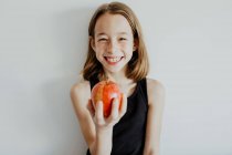 Весела дівчина в повсякденному верхньому посміхається, кусаючи свіже стигле червоне яблуко на білому тлі — стокове фото