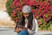 Черная женщина в повседневной одежде сидит в городском парке и болтает в социальных сетях через смартфон — стоковое фото