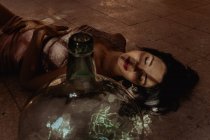 Von oben von sanftem Weibchen mit geschlossenen Augen und im Kleid auf dem Boden liegend neben Glasflasche im Raum mit Sonnenlicht — Stockfoto