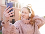 Selbstbewusste junge Frau in warmem Pullover und Kopfhörer berührt lange wellige Haare, während sie an sonnigen Tagen in der Stadt ein Selfie mit dem Smartphone macht — Stockfoto