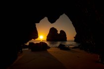 Através de caverna rochosa de pitoresca praia de areia do oceano contra o céu do pôr-do-sol na Praia do Camilo no Algarve, Portugal — Fotografia de Stock