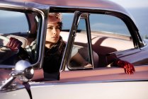 Трендова модна жінка з квіткою на капелюсі і шкіряними рукавичками, що торкаються керма в автомобілі, дивлячись вбік — стокове фото