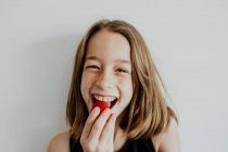 Encantado adolescente menina sorrindo e olhando para a câmera enquanto mordendo saboroso morango doce contra fundo branco — Fotografia de Stock
