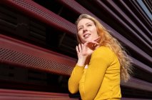Vue latérale d'une jeune femme millénaire dans des vêtements décontractés avec de longs cheveux blonds ondulés debout près d'une clôture en métal rouge — Photo de stock