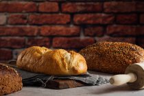 Pão branco e centeio com cereais e crosta apetitosa na tábua de corte contra a parede de tijolos na padaria — Fotografia de Stock