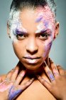 Kreative ethnische weibliche Modell mit Gesicht verschmiert mit rosa und weißer Farbe berühren Hals Blick auf Kamera auf grauem Hintergrund im Studio — Stockfoto