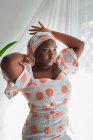 Сконцентрована молода африканка в стильному літньому одязі, регулює традиційний тюрбан, стоячи біля вікна у світлій кімнаті. — стокове фото