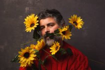 Kreativer reifer Mann im Strickpullover, der das Gesicht mit hellen Sonnenblumen vor schwarzem Hintergrund bedeckt — Stockfoto