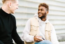 Side view of happy young multiracial male friends in trendy outfits standing on city street near modern building — Stock Photo