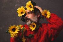 Creative mature male in knitted sweater covering face with bright sunflowers against black background — Stock Photo