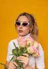 Joven hermosa hembra en traje blanco y gafas de sol de moda sosteniendo delicadas rosas rosadas mientras está de pie sobre fondo amarillo en el estudio de fotos - foto de stock