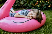 Маленький мальчик в повседневной одежде лежит на надувном розовом фламинго, веселясь на травянистой лужайке в парке — стоковое фото