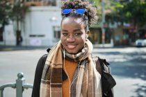 Jeune femme afro-américaine heureuse à la mode avec sac à dos et lunettes de soleil se promenant sur la chaussée urbaine tout en regardant la caméra en plein soleil — Photo de stock