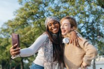 Mulher tomando selfie de amigo feminino branco enquanto relaxa no parque na cidade — Fotografia de Stock