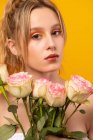 Молодая неэмоциональная красивая женщина в белом платье с голыми плечами, держа тонкие розовые розы, стоя, глядя на камеру на желтом фоне в фотостудии — стоковое фото