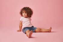 Jolie fille réfléchie tout-petit avec des cheveux bouclés dans un vêtement décontracté regardant loin tout en étant assis sur fond rose — Photo de stock