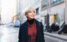 Attractive positive female in trendy wear and warm coat standing with hands in pockets on snowy city street and looking away on clear winter day in Madrid, Spain — Stock Photo