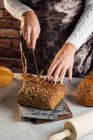 Crop anonimo panettiere femminile con coltello taglio pagnotta di pane fresco con semi di girasole sul tavolo in panetteria — Foto stock