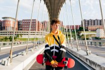 Beau garçon caucasien avec planche à roulettes debout au milieu du pont — Photo de stock