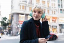 Happy young female in warm coat listening to favorite song via wireless headphones and browsing mobile phone while standing on snowy city street and looking away with toothy smile in Madrid, Spain — Stock Photo