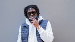 Young African American male with beard and dreadlocks smoking cigarette standing on gray wall looking at camera — Stock Photo
