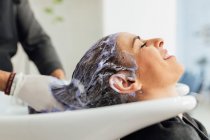 Cultiver coiffeur masculin dans le tablier laver les cheveux du client féminin dans l'évier après la coupe et la teinture dans le salon de beauté moderne — Photo de stock