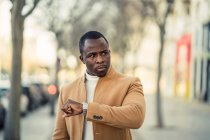 Concentrated young African American male in trendy clothes checking time on wristwatch while walking on city street on sunny day — Stock Photo