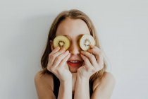 Menina alegre em roupas casuais sorrindo enquanto cobre os olhos com fatias de kiwi contra fundo branco — Fotografia de Stock