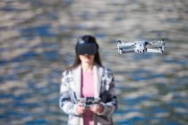 Excited female wearing modern VR goggles operating drone with remote controller and experiencing virtual reality while standing against blurred sea — Stock Photo