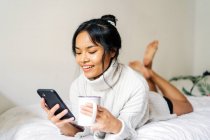 Lange Haare brünett asiatische Frau liegend auf dem Bett zu Hause und mit einem Handy — Stockfoto