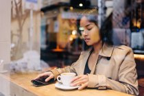 Довговолоса брюнетка азіатка, яка п'є каву на кав'ярні, дивлячись на мобільний телефон. — стокове фото