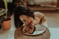 Peaceful woman sitting with closed eyes on floor in room and reflecting in round shaped mirror — Stock Photo