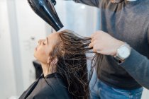 Растениеводство молодой этнический мужчина парикмахер сушки волос клиентки с закрытыми глазами в современной студии красоты — стоковое фото