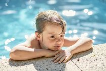 Desde arriba de un lindo niño reflexivo apoyado en el borde de la piscina mientras descansa después de nadar en un día soleado - foto de stock