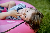 Cheerful little boy in casual clothes lying on inflatable pink flamingo while having fun on grassy lawn in park — Stock Photo