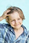 Adorable little boy with blond hair in stylish checkered shirt smiling and looking away while standing against blue background in sunlight — Stock Photo