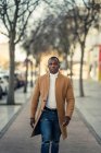 Confiante jovem étnico masculino na roupa da moda andando na rua da cidade e olhando para a câmera no dia ensolarado — Fotografia de Stock
