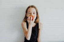 Весела дівчина в повсякденному верхньому посміхається, кусаючи свіже стигле червоне яблуко на білому тлі — стокове фото