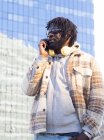 Тендітний афроамериканець у сонцезахисних окулярах з навушниками навколо шиї говорить по мобільному телефону стоячи з рукою в кишені проти будівництва, що відбиває місто. — стокове фото