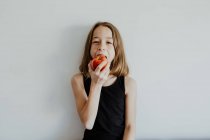 Menina pré-adolescente alegre no topo casual sorrindo enquanto morde maçã vermelha madura fresca contra fundo branco — Fotografia de Stock
