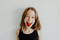 Deliziosa ragazza adolescente sorridente e guardando la fotocamera mentre morde gustosa fragola dolce contro sfondo bianco — Foto stock