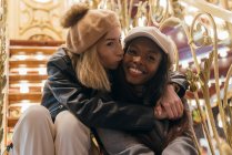 Delighted woman kissing African American female friend in cheek while enjoying weekend in amusement park in evening — Stock Photo