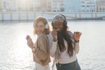 Seitenansicht von multirassischen Freundinnen mit Kopfhörern, die an sonnigen Tagen gemeinsam Musik genießen und auf einem Damm tanzen — Stockfoto
