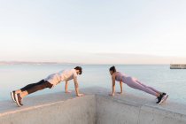 Side view of anonymous young sportsman with ethnic female athlete in active wear working out on embankment against ocean — Stock Photo