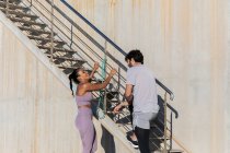 Seitenansicht eines jungen Sportlers mit überkreuzten Beinen, der mit einer lächelnden Partnerin in Sportbekleidung spricht, während er sich in der Nähe der Treppe anschaut — Stockfoto