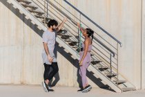 Side view of young sportsman with crossed legs speaking with smiling female partner in sportswear while looking at each other near staircase — Stock Photo