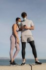 Невпізнавана етнічна спортсменка в стерильній масці поблизу партнера-чоловіка, який серфінгує в Інтернеті на мобільному телефоні проти зрізаного моря під хмарним небом — стокове фото