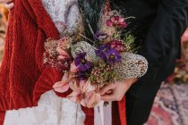 Mariée méconnaissable et marié portant des tenues de mariage traditionnelles avec des fleurs colorées dans les mains debout sur le tapis dans la nature pendant la célébration du mariage — Photo de stock