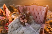 Fauteuil rose avec des fleurs sur plaid moelleux placé sur un tapis vintage avec des plumes décoratives au sommet de la colline dans la nature lors de la célébration du mariage — Photo de stock